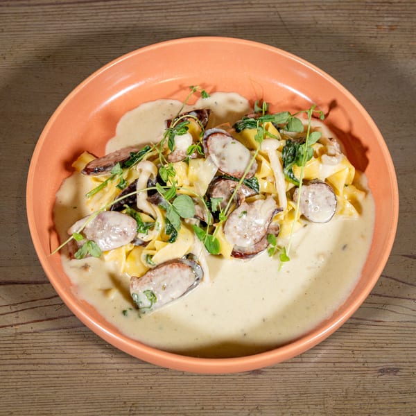 Creamy pasta with sliced sausage, mushrooms, and fresh herbs in an orange bowl on a wooden table.