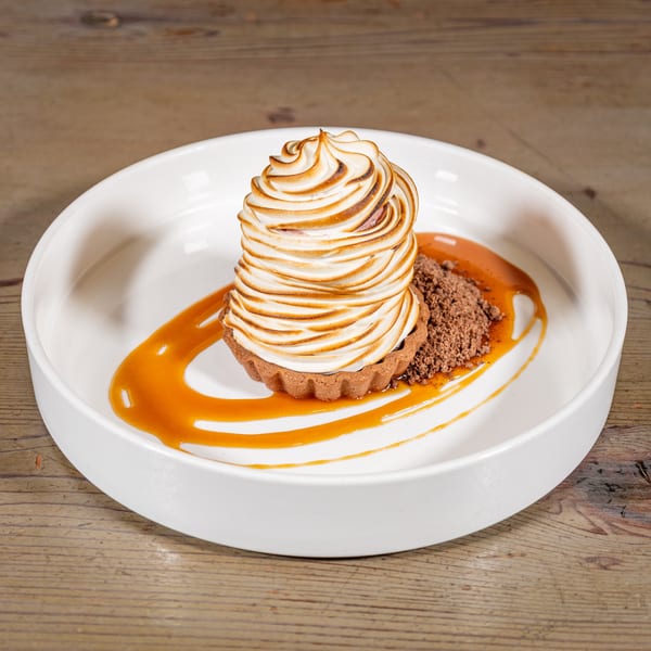 A dessert tart with toasted meringue, chocolate crumbles, and caramel sauce in a white bowl.