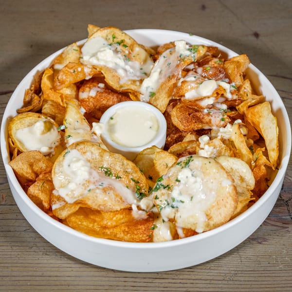 A white bowl filled with crispy potato chips, drizzled with creamy white sauce and herbs.