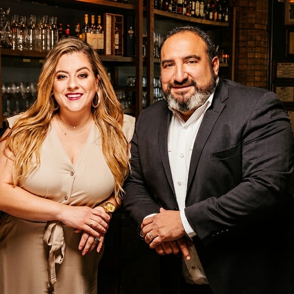 Two restaurant owners, a woman and a man, smiling in front of a bar with liquor bottles.