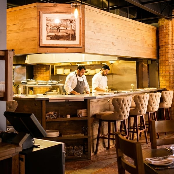 Two chefs working in an open kitchen with bar seating and dining tables in a restaurant.