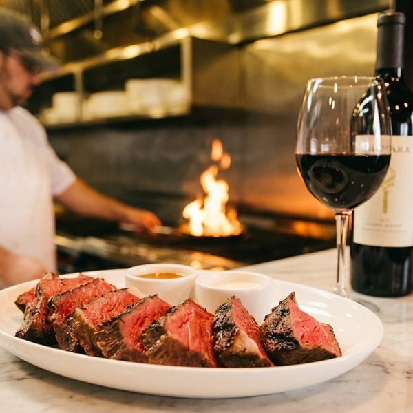 Sliced steak with sauces and red wine on a counter, with a chef cooking over flames in the background.