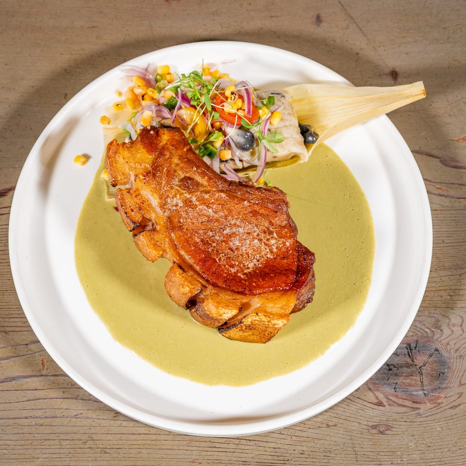 Crispy pork belly in green sauce with a tamale and corn salsa on a white plate.