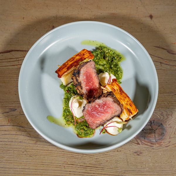 Grilled steak slices with green pesto, white sauce, and crispy potato gratin on a blue plate.