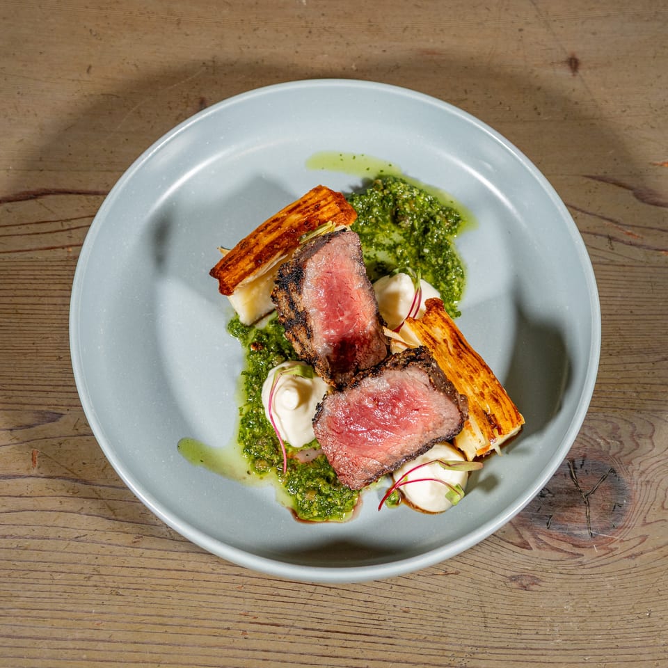 Seared steak with green pesto, white cream, and crispy potato gratin on a light blue plate.