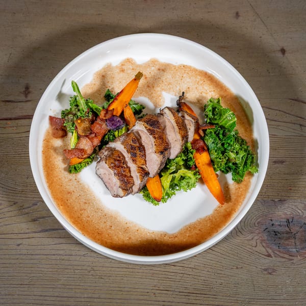 Sliced duck breast with roasted carrots, kale, and bacon on a white plate with sauce.