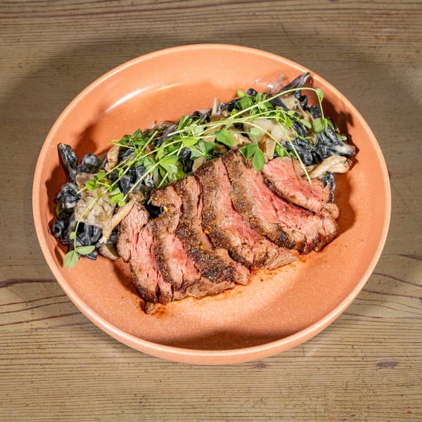 Sliced steak with black pasta, mushrooms, and microgreens on an orange plate.